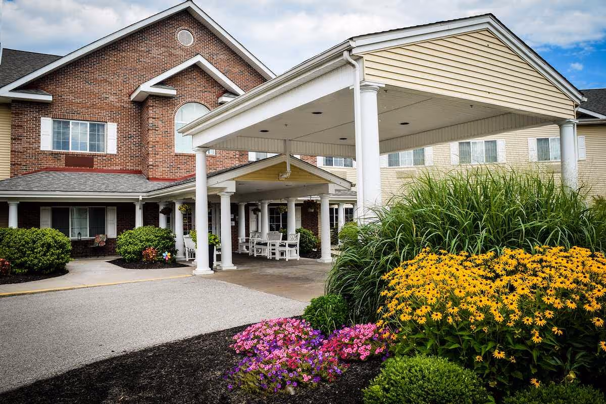Entrance of a senior living facility with a covered driveway supported by white columns. The building features a combination of red brick and beige siding with multiple windows. There are well-maintained flower beds with yellow and purple flowers and green shrubs near the entrance.