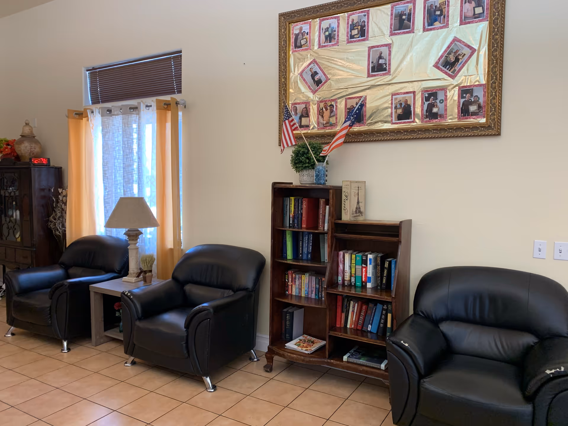 A cozy sitting area with three black leather armchairs arranged along a wall. Between two of the chairs is a small wooden side table with a lamp and a small plant. Behind the chairs is a window with yellow curtains and a brown blind. To the right is a wooden bookshelf filled with books and decorative items, topped with two small American flags and a plant. Above the bookshelf is a large framed collage of photographs. The floor is tiled and the walls are painted light beige.