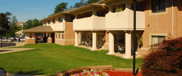 Exterior view of a two-story senior living facility building with balconies and a covered entrance. There is a well-maintained lawn with a flower bed and some shrubs in the foreground, and a parking area with cars and people in the background.