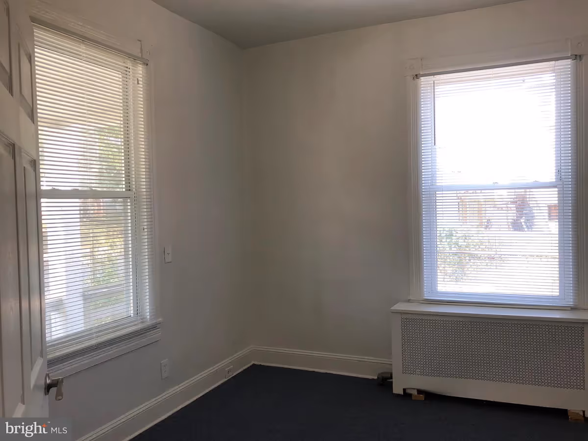 Empty room with two windows covered by white blinds, white walls, a white radiator cover beneath one window, and a dark carpeted floor.
