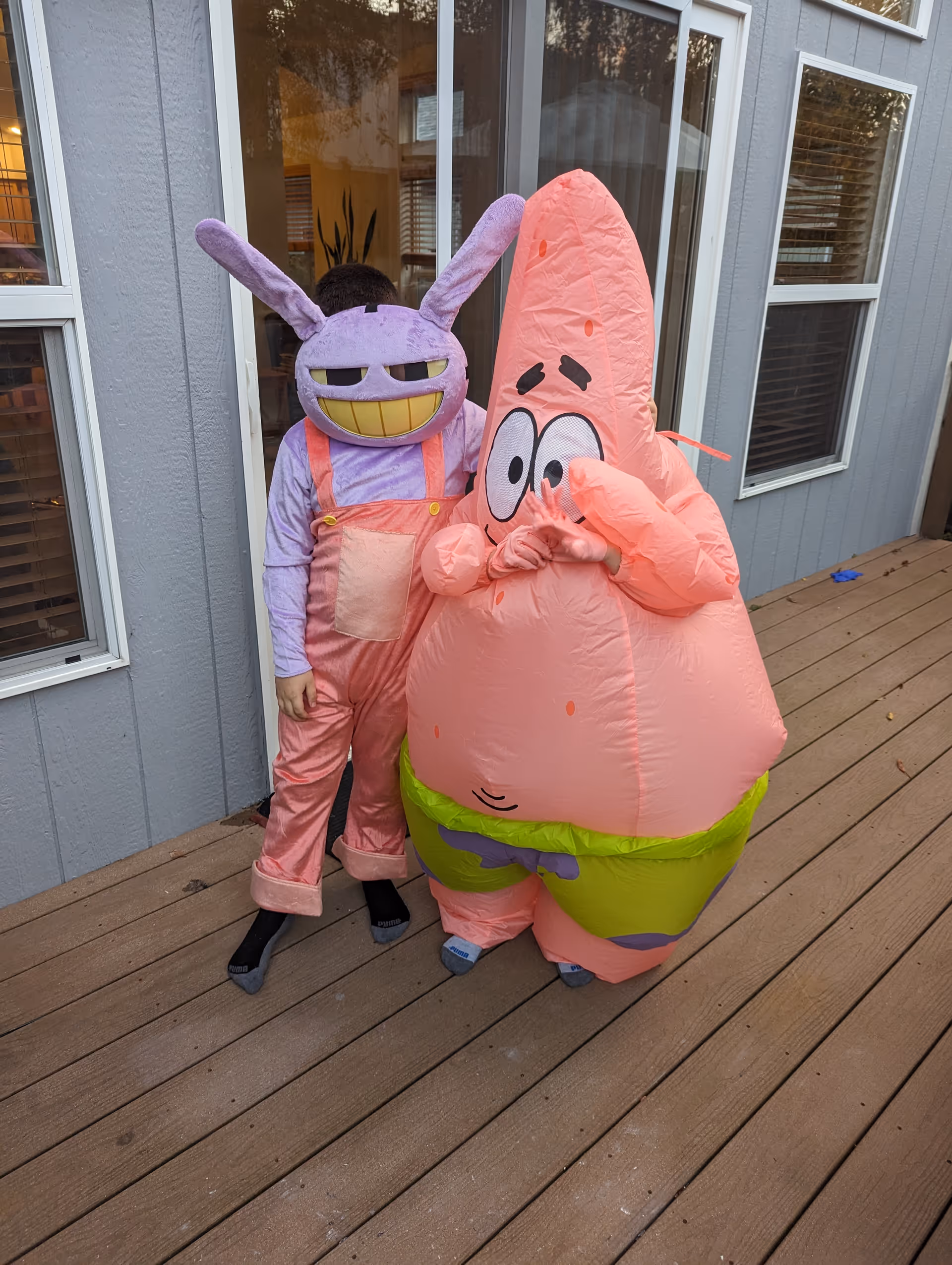 Two children standing on a wooden deck outside a gray house with white-trimmed windows. One child is wearing a purple bunny mask with a wide grin and pink overalls, while the other child is dressed in an inflatable Patrick Star costume from SpongeBob SquarePants.