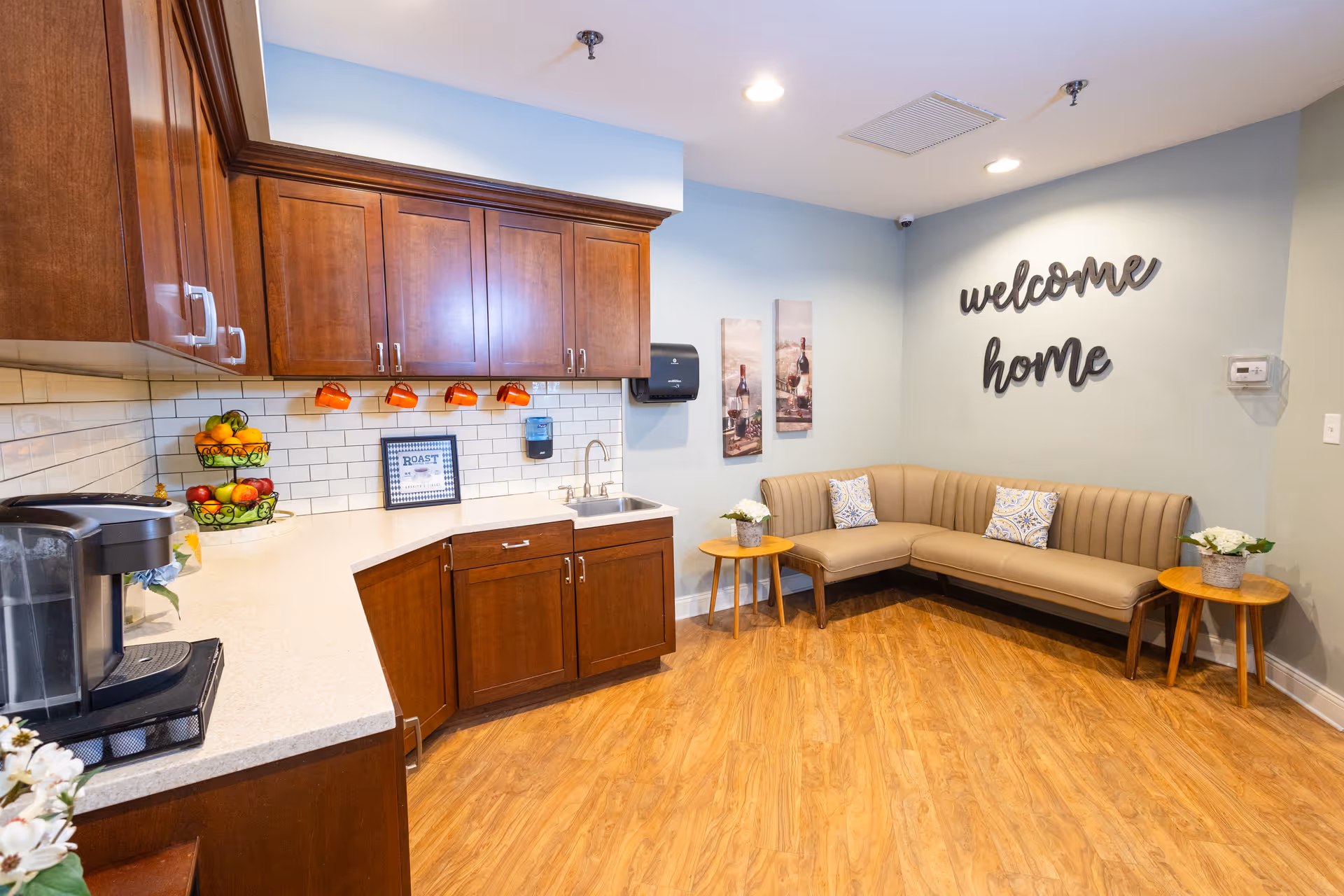 A bright common kitchenette and lounge with wooden cabinets, a coffee station and an L-shaped beige bench under a 'welcome home' wall sign.