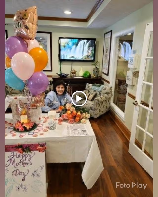 A woman sitting at a table decorated with flowers and balloons in a cozy living room. The table has a 'Happy Mothers Day' sign, and the room features floral-patterned armchairs, a wooden floor, framed artwork, and a wall-mounted TV displaying a waterfall scene.