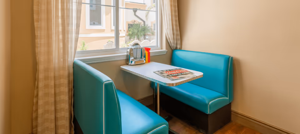 A cozy dining nook with turquoise cushioned bench seats on either side of a small rectangular table. On the table, there is a vintage-style jukebox, a ketchup bottle, and a Coca-Cola menu. The nook is next to a window with beige curtains, allowing natural light to brighten the space.