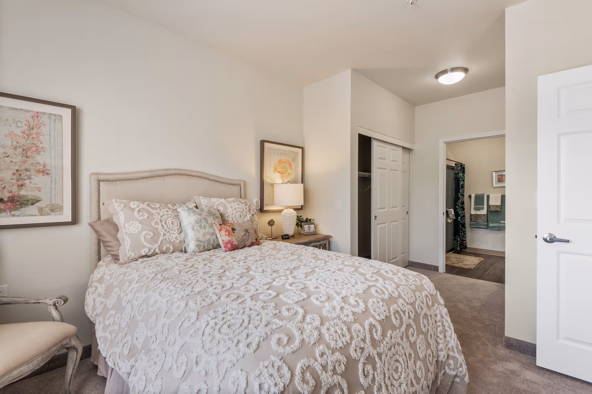A cozy bedroom with a beige upholstered bed covered in a textured white and beige bedspread. The bed has multiple decorative pillows. There is a wooden nightstand with a white lamp and framed pictures. A framed floral artwork hangs on the wall above the nightstand. To the right, there is a closet with sliding doors and an open door leading to a bathroom with towels hanging on the wall and a shower curtain with a floral pattern. A cushioned chair is partially visible on the left side of the image.