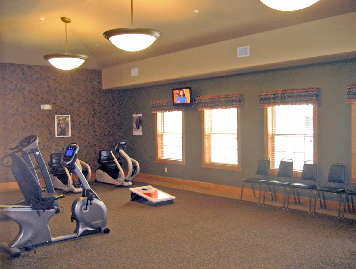 A senior community exercise room with recumbent bikes, a cornhole board, chairs along windows, and ceiling lights.