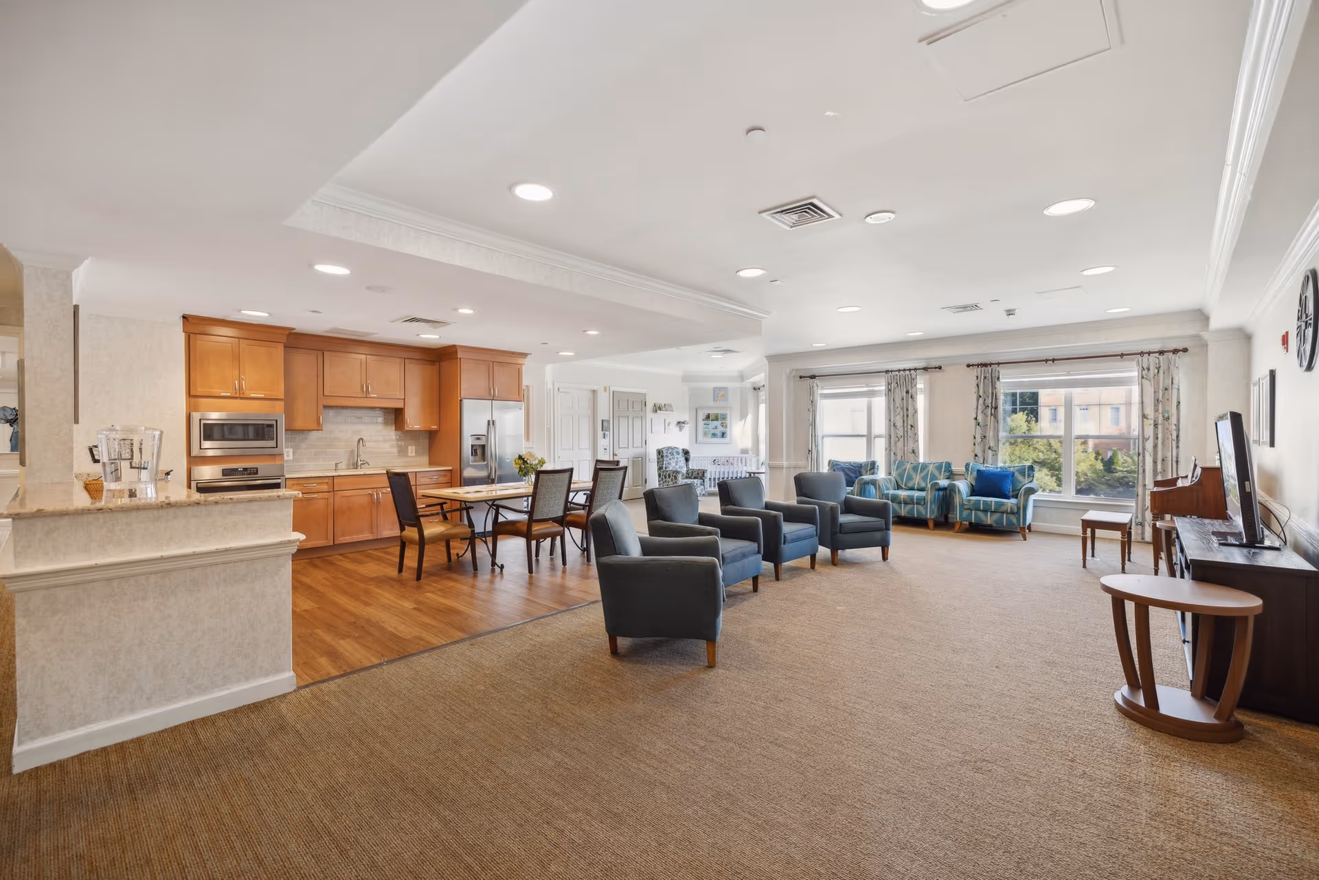 A spacious and well-lit common area in a senior living facility featuring a kitchen with wooden cabinets, stainless steel appliances, and a dining table with chairs. Adjacent to the kitchen is a living room area with multiple armchairs and sofas arranged around a large window with curtains, allowing natural light to fill the room. The space has a carpeted floor and recessed ceiling lights.