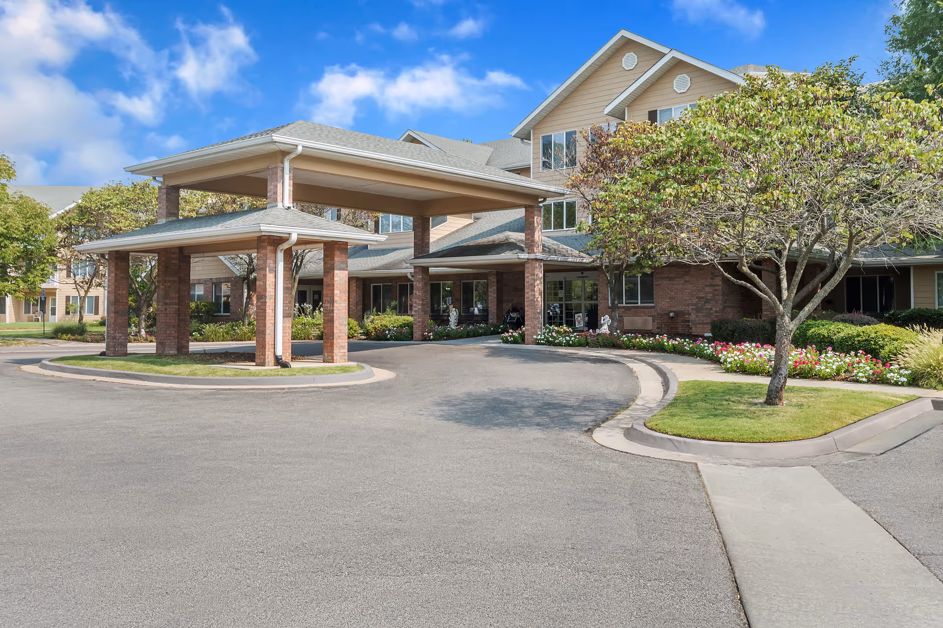 Exterior view of Morada Broken Arrow senior living facility showing a covered entrance with brick pillars, a driveway, landscaped flower beds, and trees under a blue sky with some clouds.