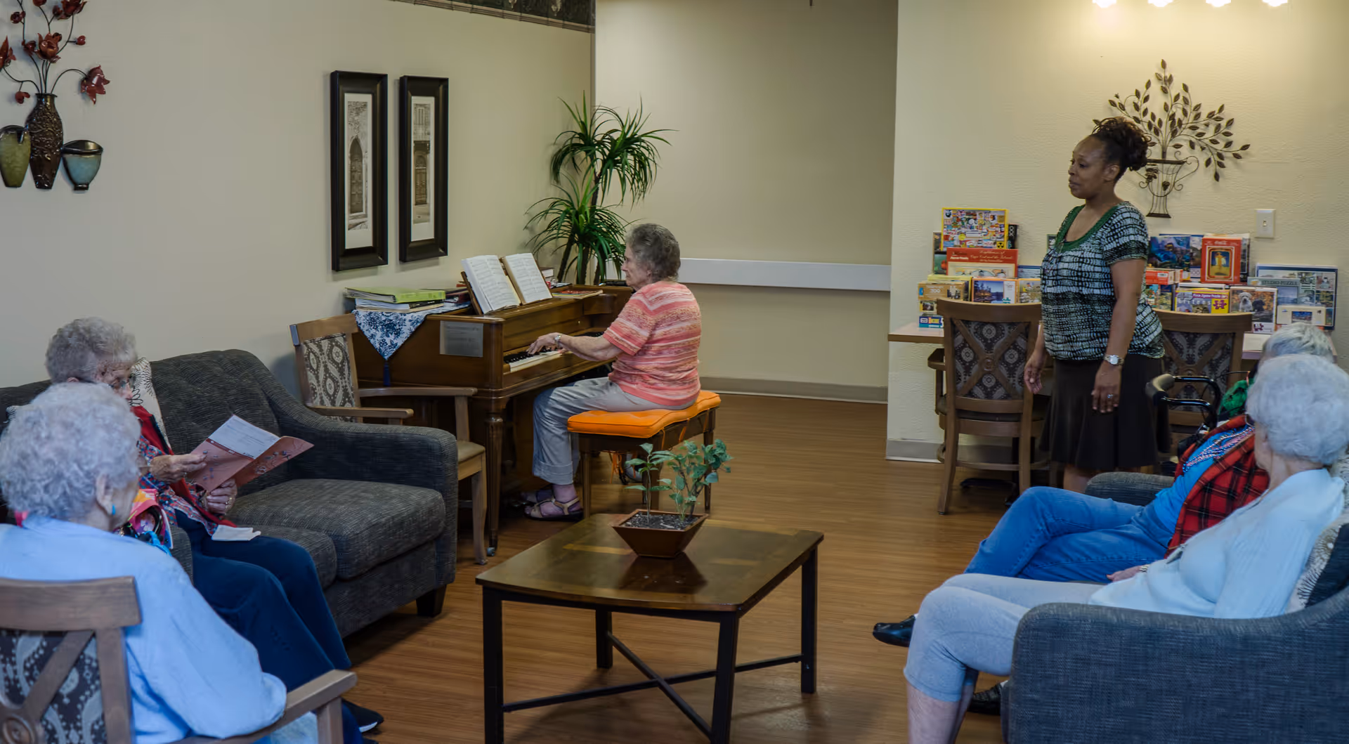 A group of elderly women seated in a living room area, listening to one woman playing a piano. Another woman stands nearby. The room has comfortable seating, a coffee table with a small plant, framed artwork on the walls, and a table with puzzles and games in the background.