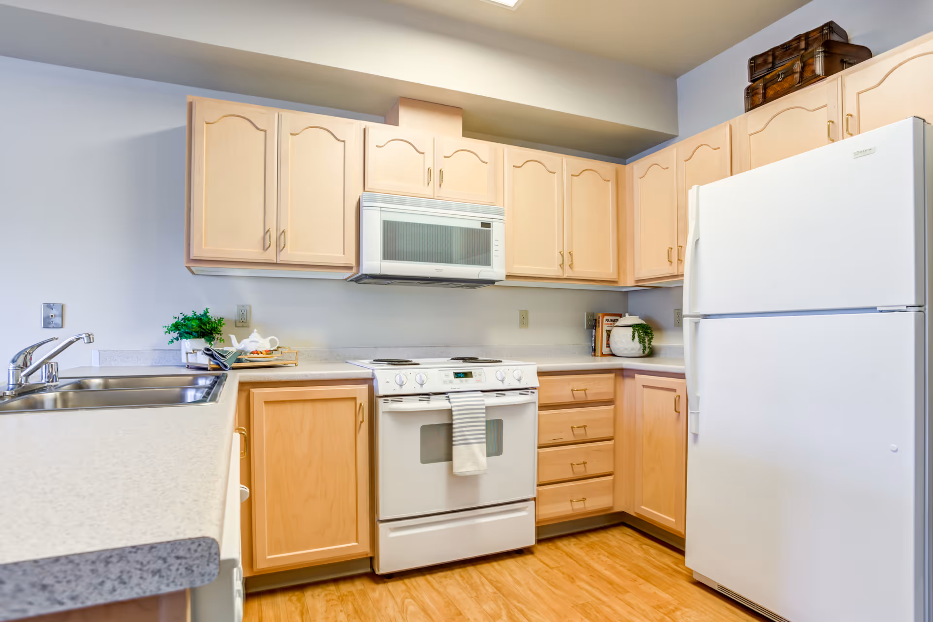 A clean and modern kitchen with light wood cabinets, a white refrigerator, a white stove with an oven, a microwave above the stove, a double sink with a chrome faucet, and a light-colored countertop. There are small decorative plants and kitchen items on the counters.