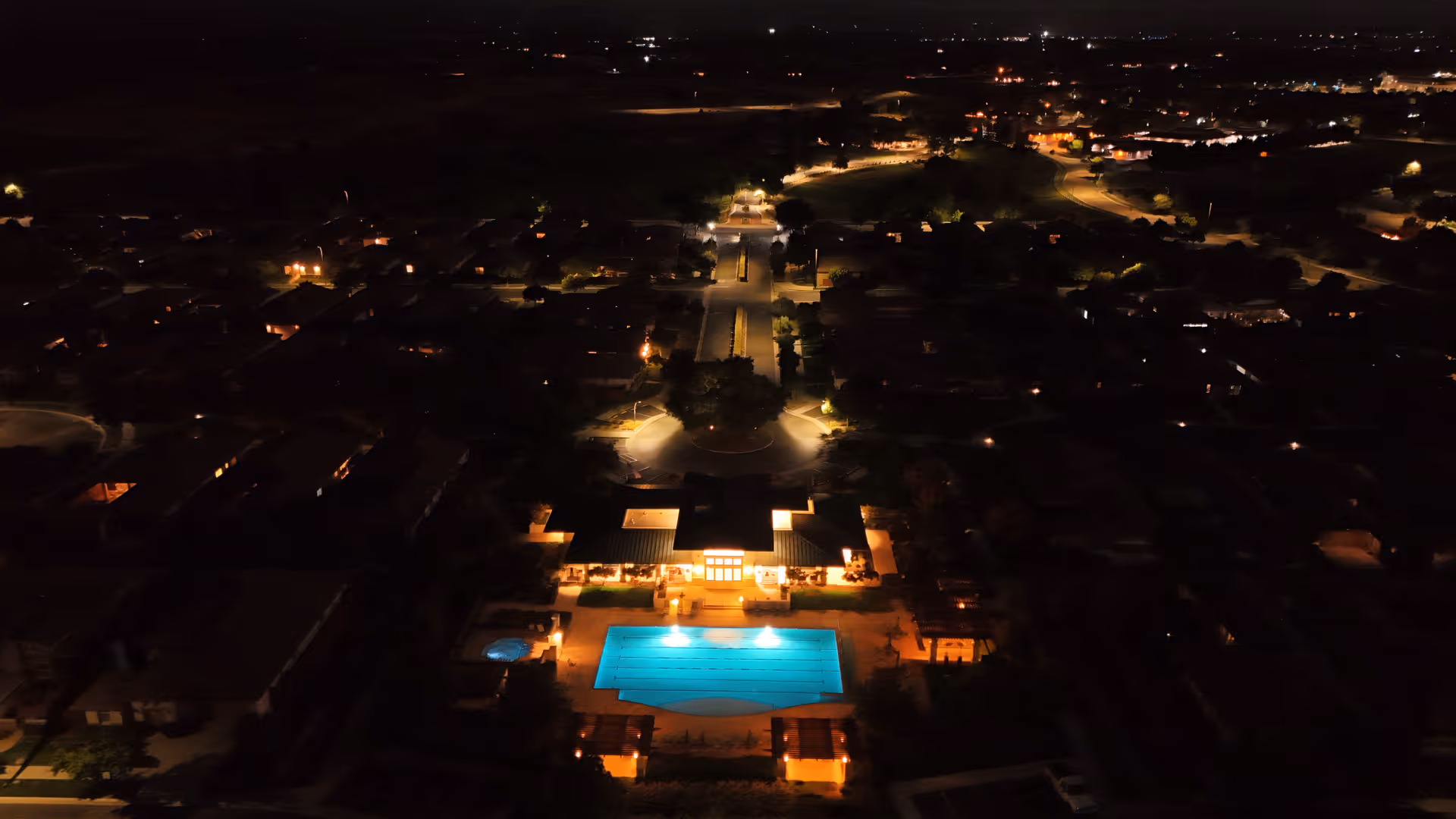 Nighttime aerial view of a lit clubhouse and swimming pool surrounded by a dark residential neighborhood.
