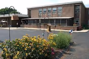 Brick senior care facility with a front entrance, large windows, and flowering foreground landscaping.