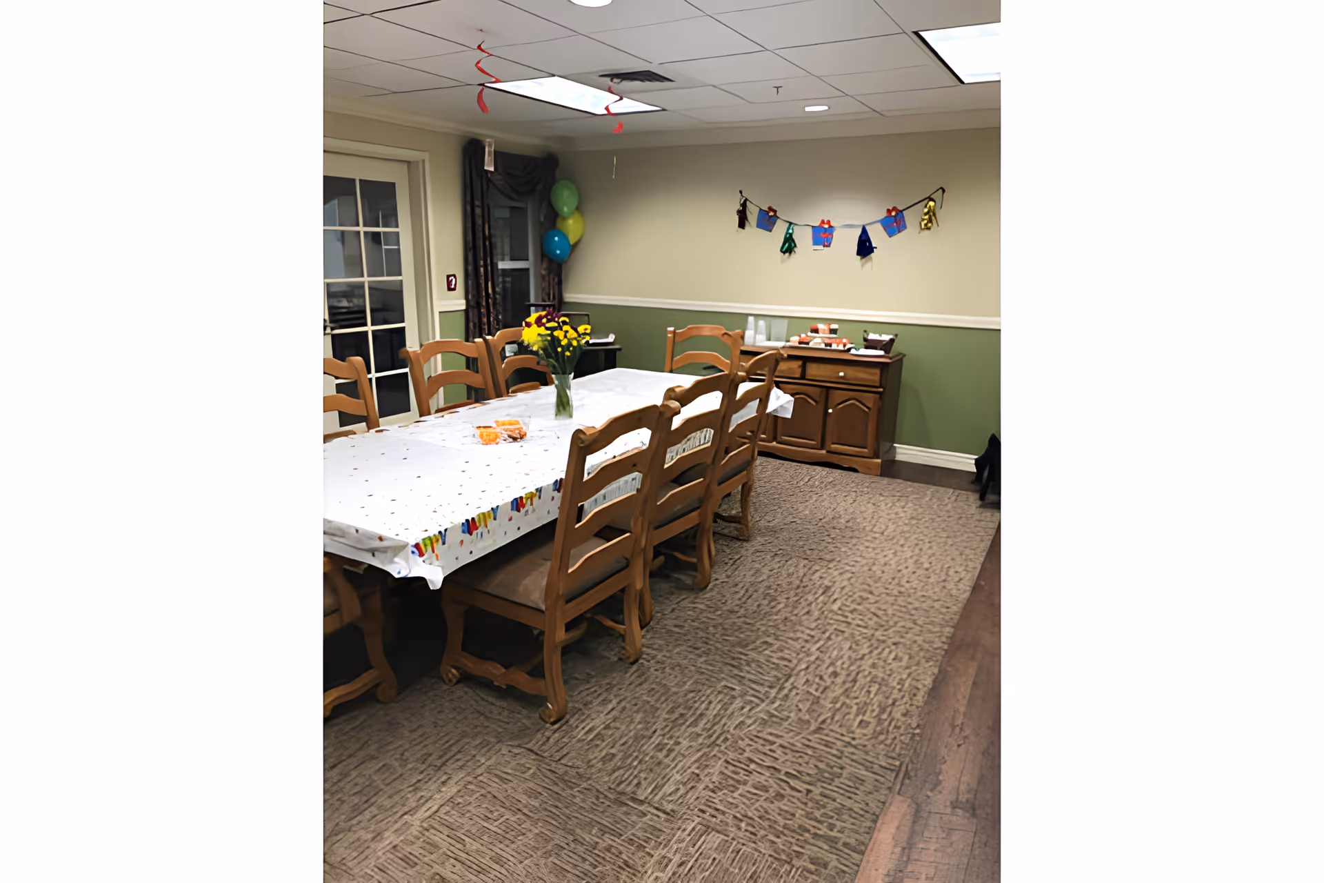 A dining room with a long wooden table covered with a white tablecloth decorated with colorful patterns. Eight wooden chairs surround the table. A vase with yellow and purple flowers is placed in the center of the table. In the background, there is a wooden sideboard with cups and snacks on top. The walls are painted green on the lower half and beige on the upper half, with a decorative garland hanging on the wall. Balloons are visible near a window with dark curtains.