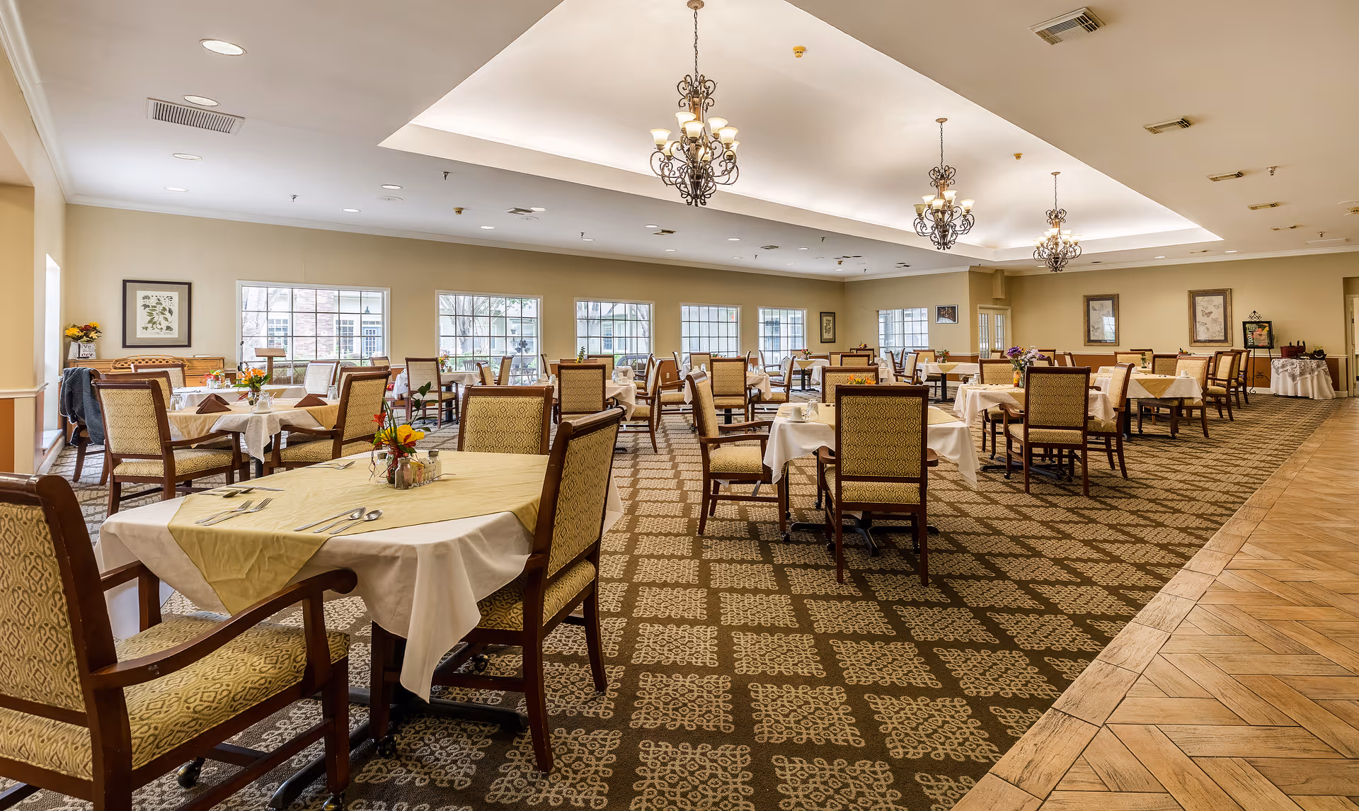 A spacious dining room with multiple tables covered with white and beige tablecloths, each set with chairs and small floral centerpieces. The room features large windows allowing natural light to fill the space, patterned carpet flooring, and elegant chandeliers hanging from the ceiling.