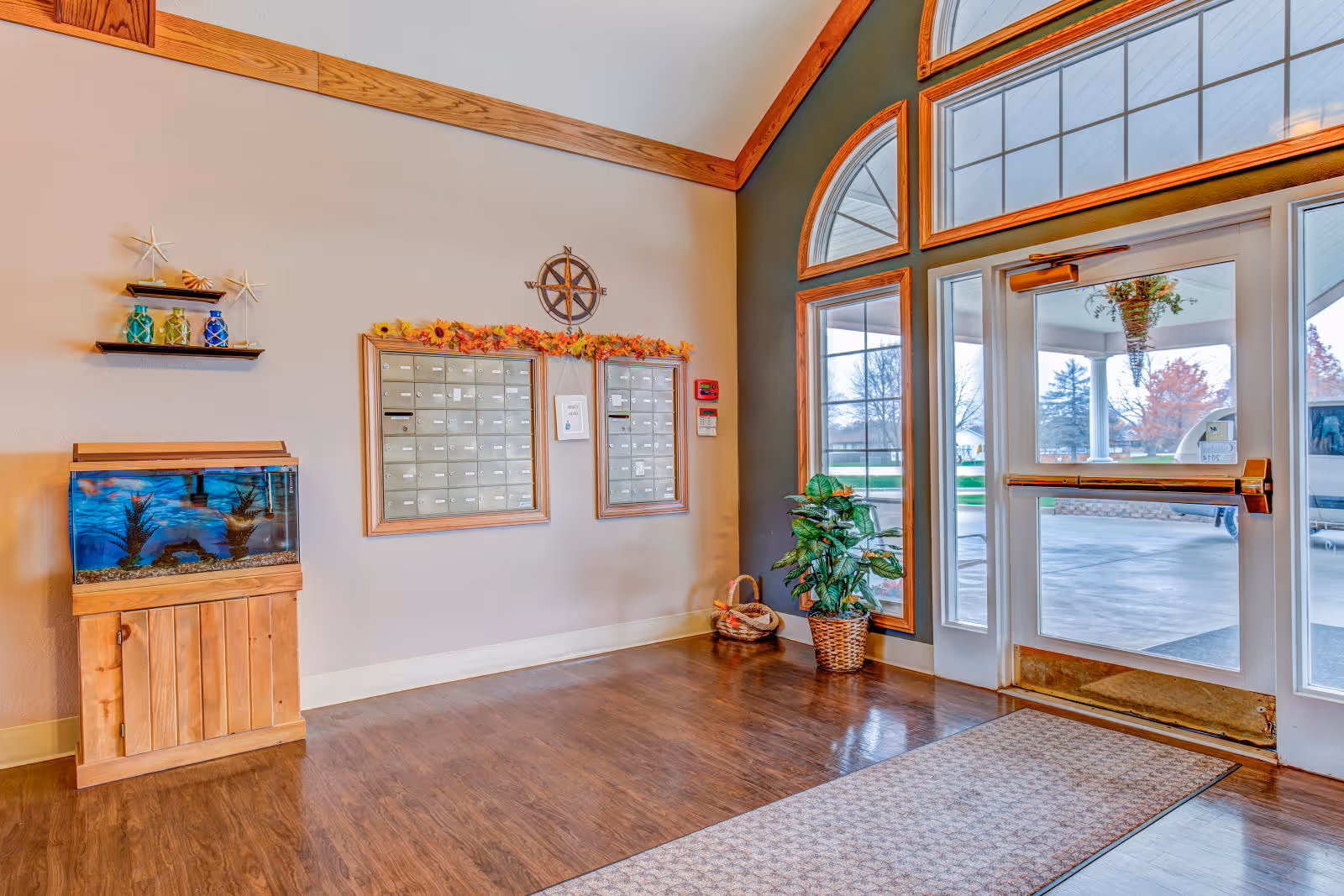 Entrance area of a senior living facility with large glass doors and windows, a wooden floor, a small fish tank on a wooden stand, two wall-mounted mailboxes decorated with a fall-themed garland, a potted plant, and decorative shelves with starfish and glass jars.