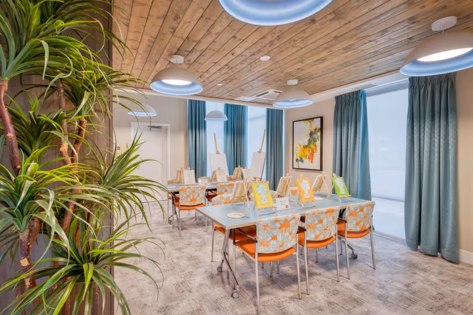 Bright interior activity room with tables set up for painting, easels, patterned chairs, large windows with teal curtains, and pendant lights.