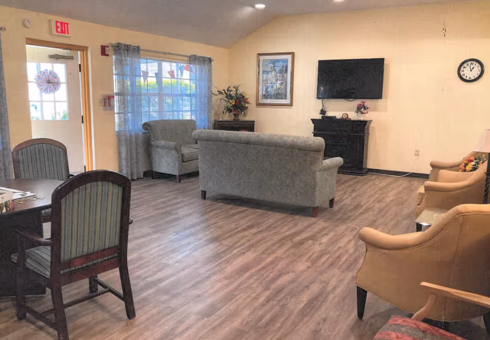 A senior living common area with sofas and chairs arranged around a wall-mounted TV and a table with chairs near a window.