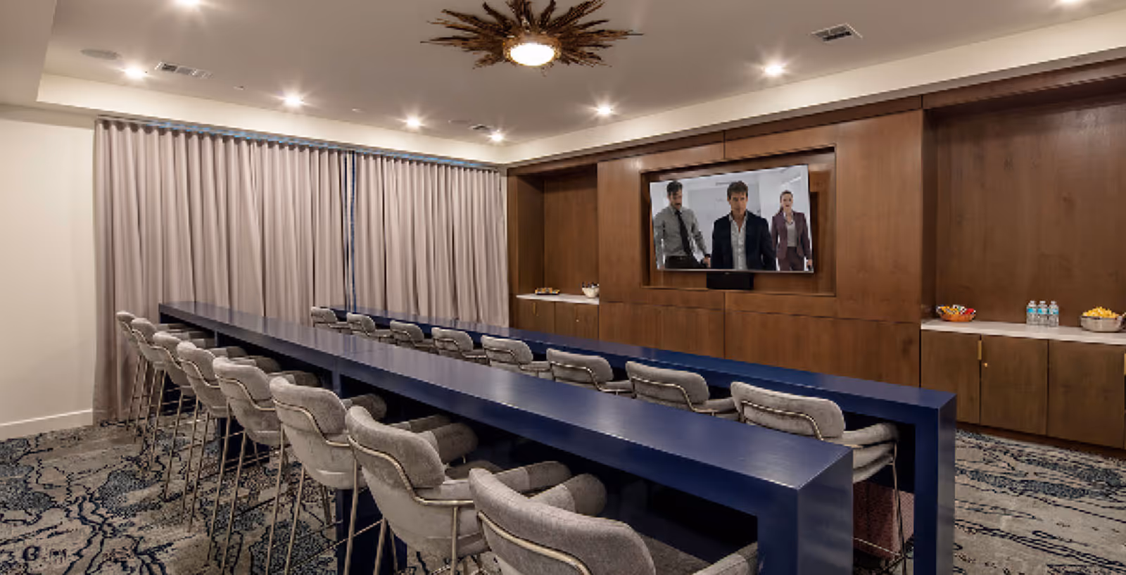 Long communal room with two navy blue bar-height tables, upholstered stools, a wall-mounted TV, and snack counters along the back.