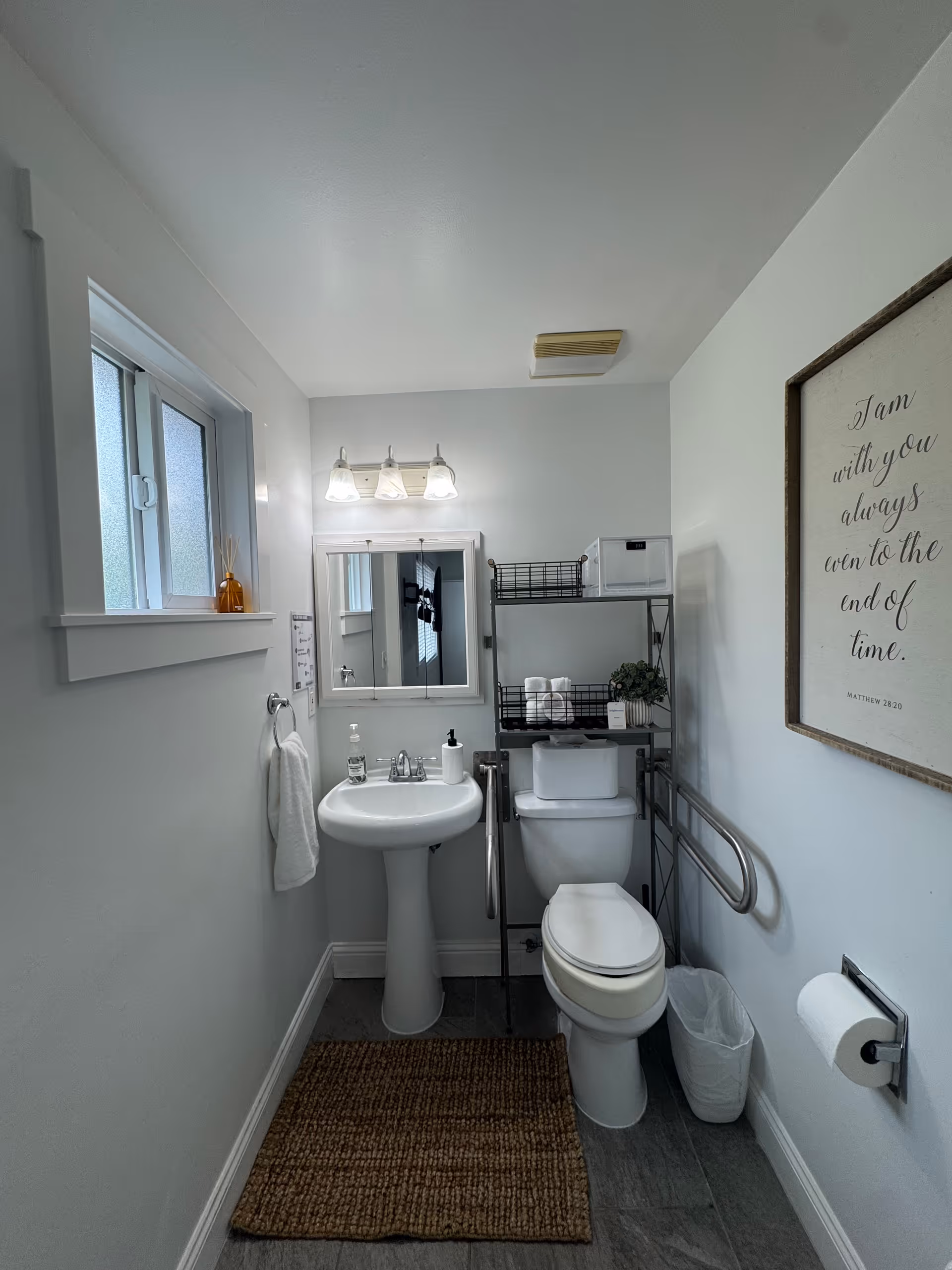 A small, clean bathroom with a white pedestal sink, a toilet with safety rails, a wall-mounted mirror with three lights above it, a small window with frosted glass, a brown woven rug on the floor, and a framed inspirational quote on the wall. There is a metal shelving unit above the toilet holding towels and a small plant.