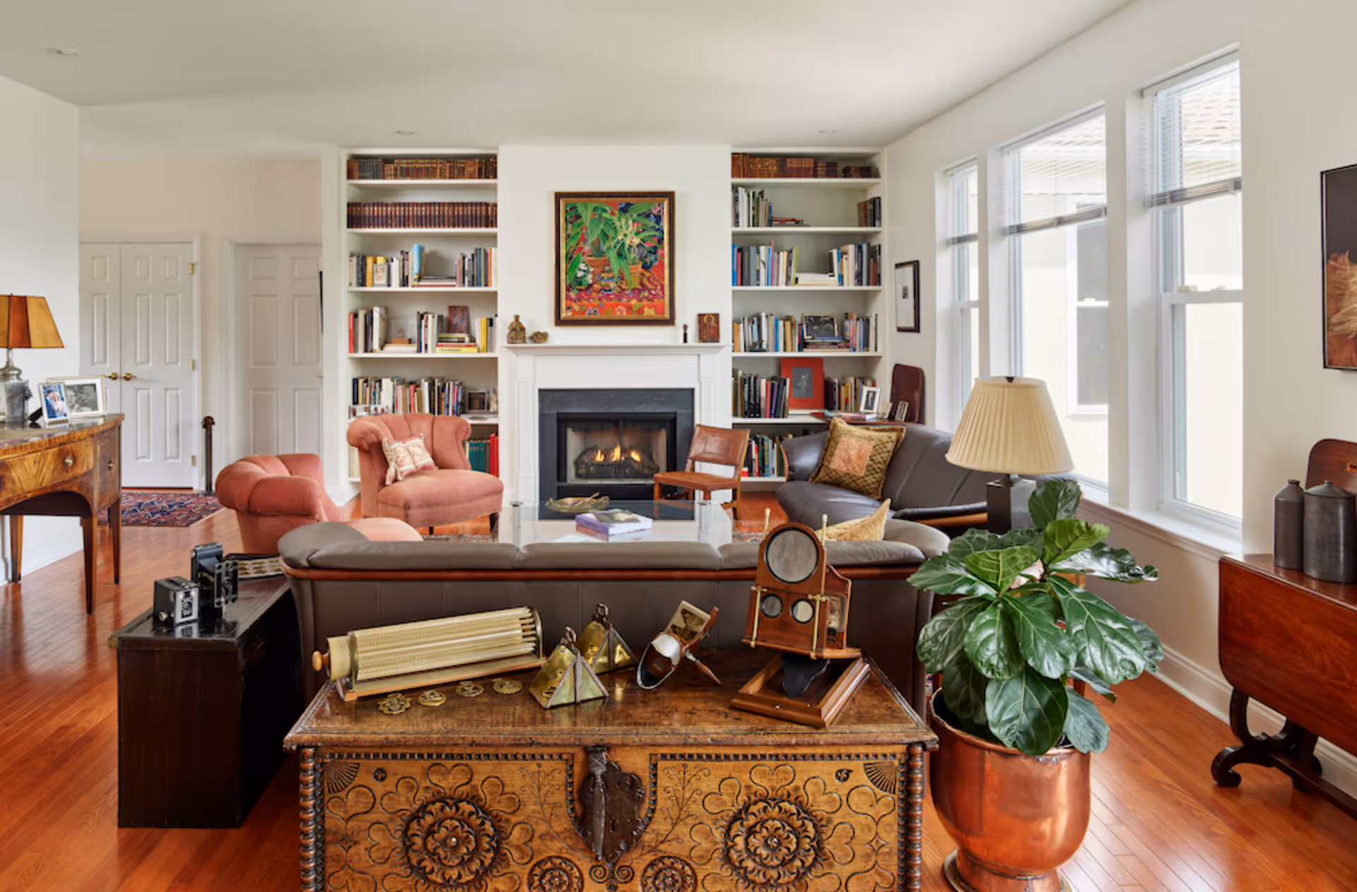 A cozy living room with a fireplace in the center, flanked by built-in bookshelves filled with books. There are two pink armchairs, a brown leather sofa, a wooden chair, and a glass coffee table. A wooden chest with decorative items sits in the foreground next to a large green potted plant in a copper container. Large windows on the right side let in natural light, and a table lamp is placed on a side table near the sofa.