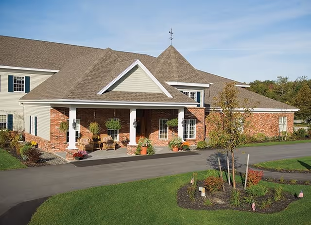 Exterior view of a senior living facility building with a covered entrance supported by white columns. The building has a combination of brick and light-colored siding with a peaked roof and a weather vane on top. There are potted plants and hanging baskets near the entrance, a paved driveway, and landscaped greenery with small trees and shrubs in the foreground under a clear blue sky.