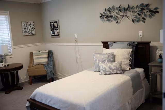 A tidy single bed in a softly lit bedroom with decorative pillows, a chair, side table, and wall art.