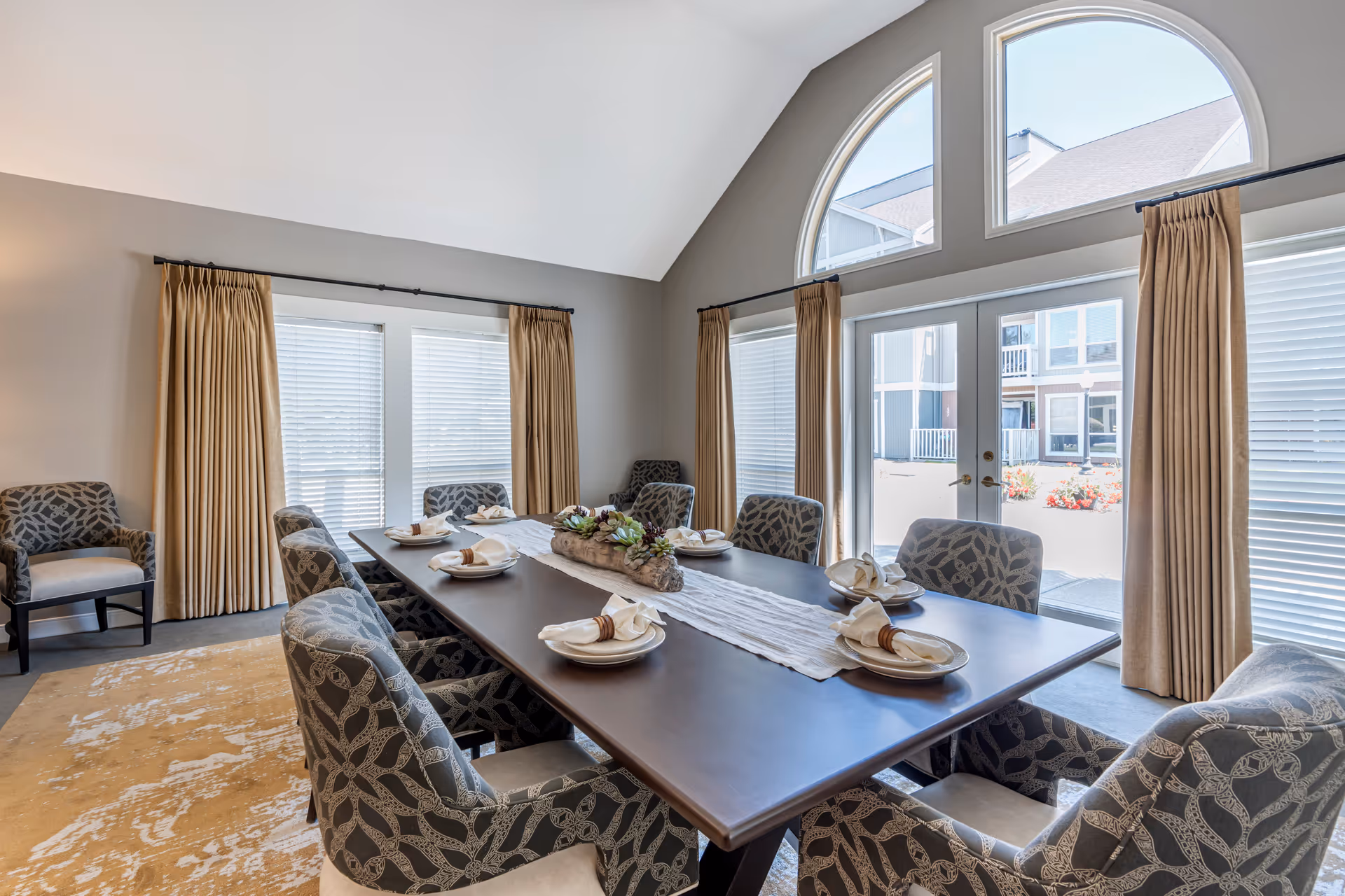 A bright dining room with a large rectangular wooden table set for eight people. The table has a white runner and a centerpiece with succulents. The room features large windows with beige curtains and an arched window above a glass door leading outside. There are patterned upholstered chairs around the table and a patterned armchair in the corner.