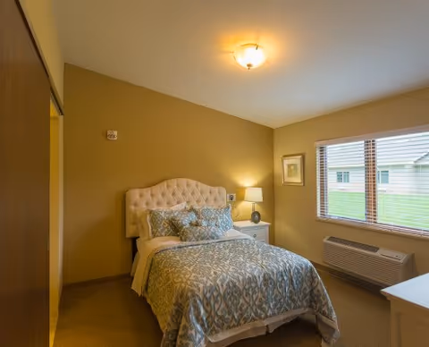 A cozy bedroom with a single bed featuring a tufted headboard and patterned bedding. There is a nightstand with a lamp and a framed picture on the wall. A window with blinds lets in natural light, and an air conditioning unit is installed below the window.