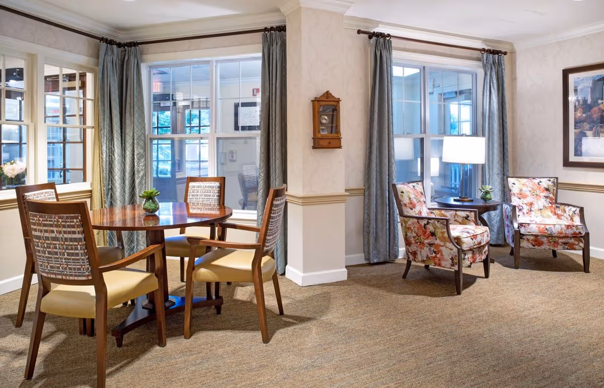 Bright communal sitting area with a round wooden dining table and chairs by large windows and two floral armchairs with a side table and lamp.