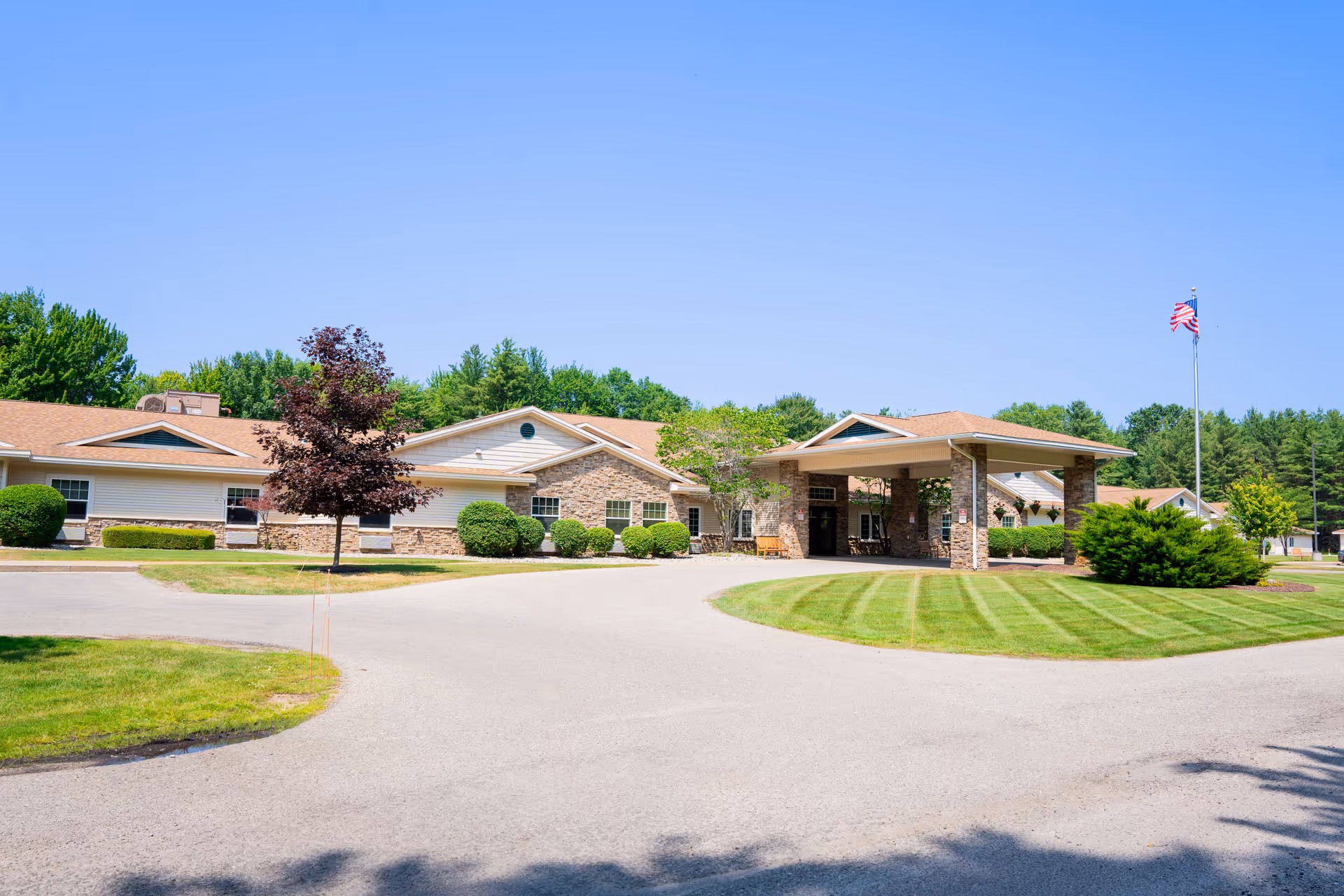 Exterior view of a single-story senior living facility building with a covered entrance, neatly trimmed bushes, a tree with red leaves, and an American flag on a flagpole under a clear blue sky.