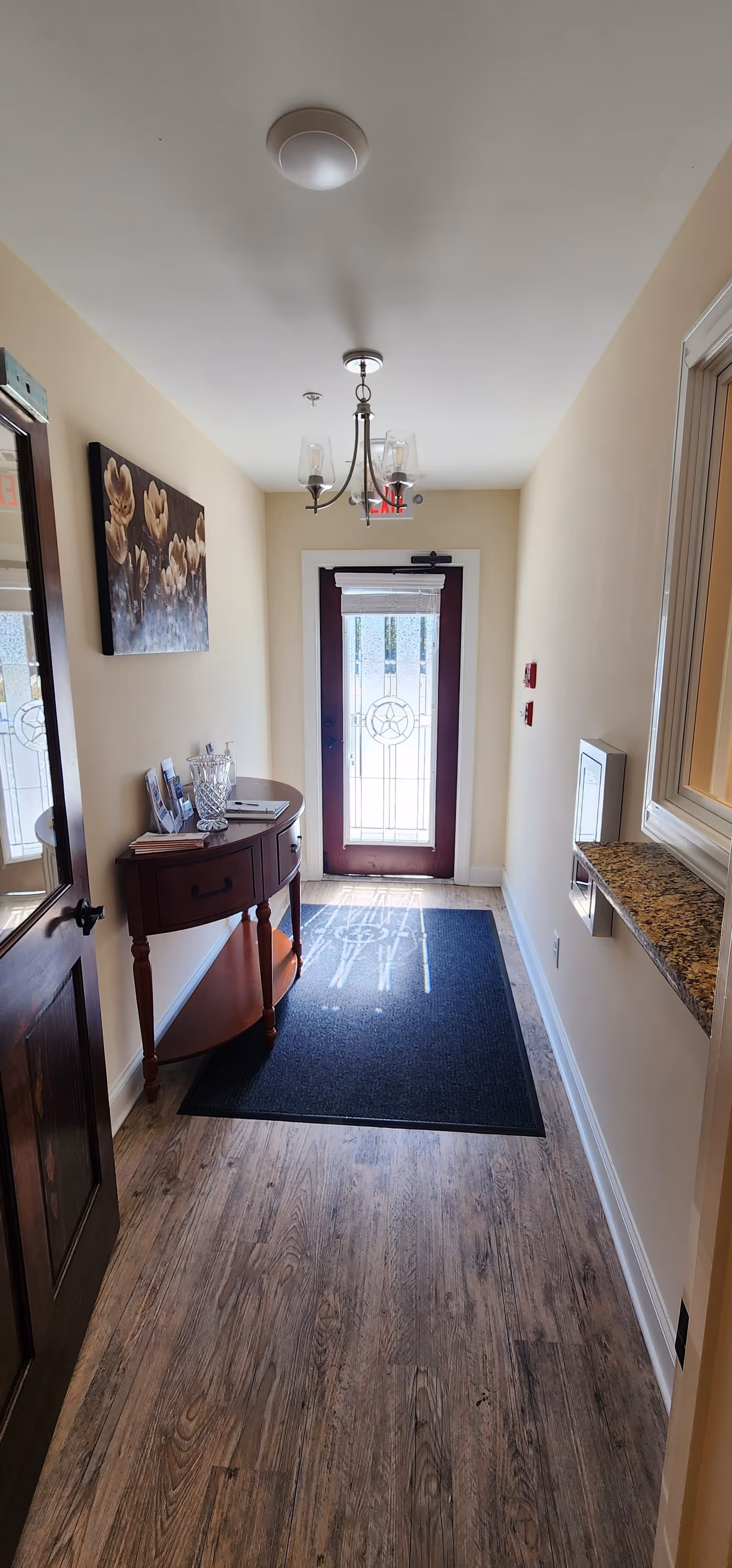 A narrow hallway with wooden flooring leading to a glass door with decorative patterns. On the left side, there is a small wooden table with a crystal vase and some framed photos, and a floral painting hangs above it. On the right side, there is a granite countertop window opening and two red fire alarm pull stations on the wall. A chandelier with three lights hangs from the ceiling.