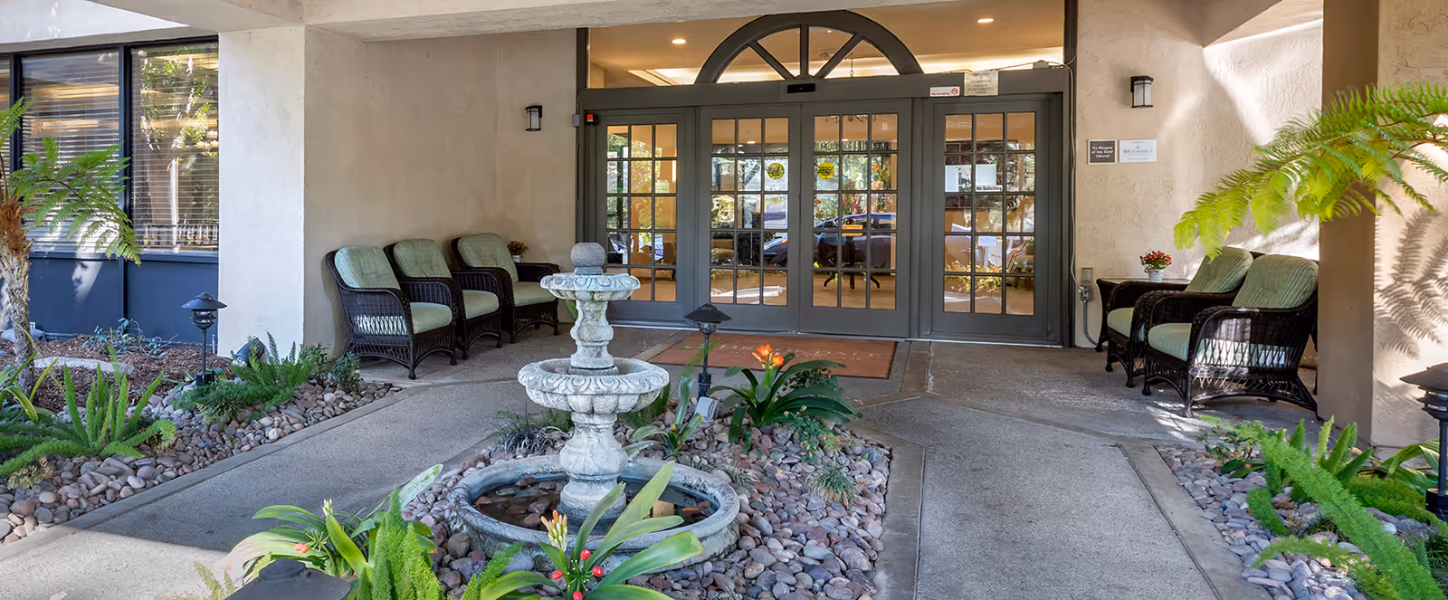 Covered entrance area of a senior living facility with a decorative stone fountain surrounded by plants and rocks in the center. There are cushioned wicker chairs arranged on both sides near the entrance doors, which are glass with multiple panes. The walls are light-colored, and there are small wall-mounted lights on either side of the entrance.