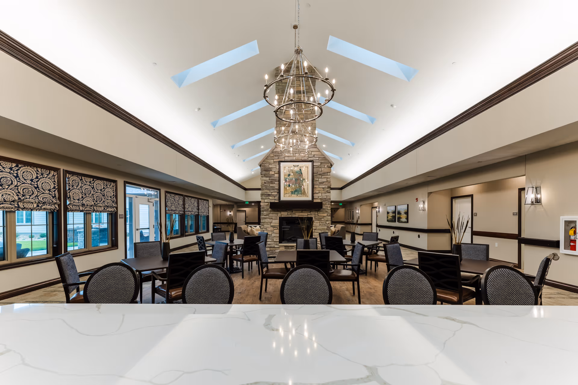 Spacious senior living dining room with multiple tables and chairs, a central stone fireplace, chandeliers, and skylights.