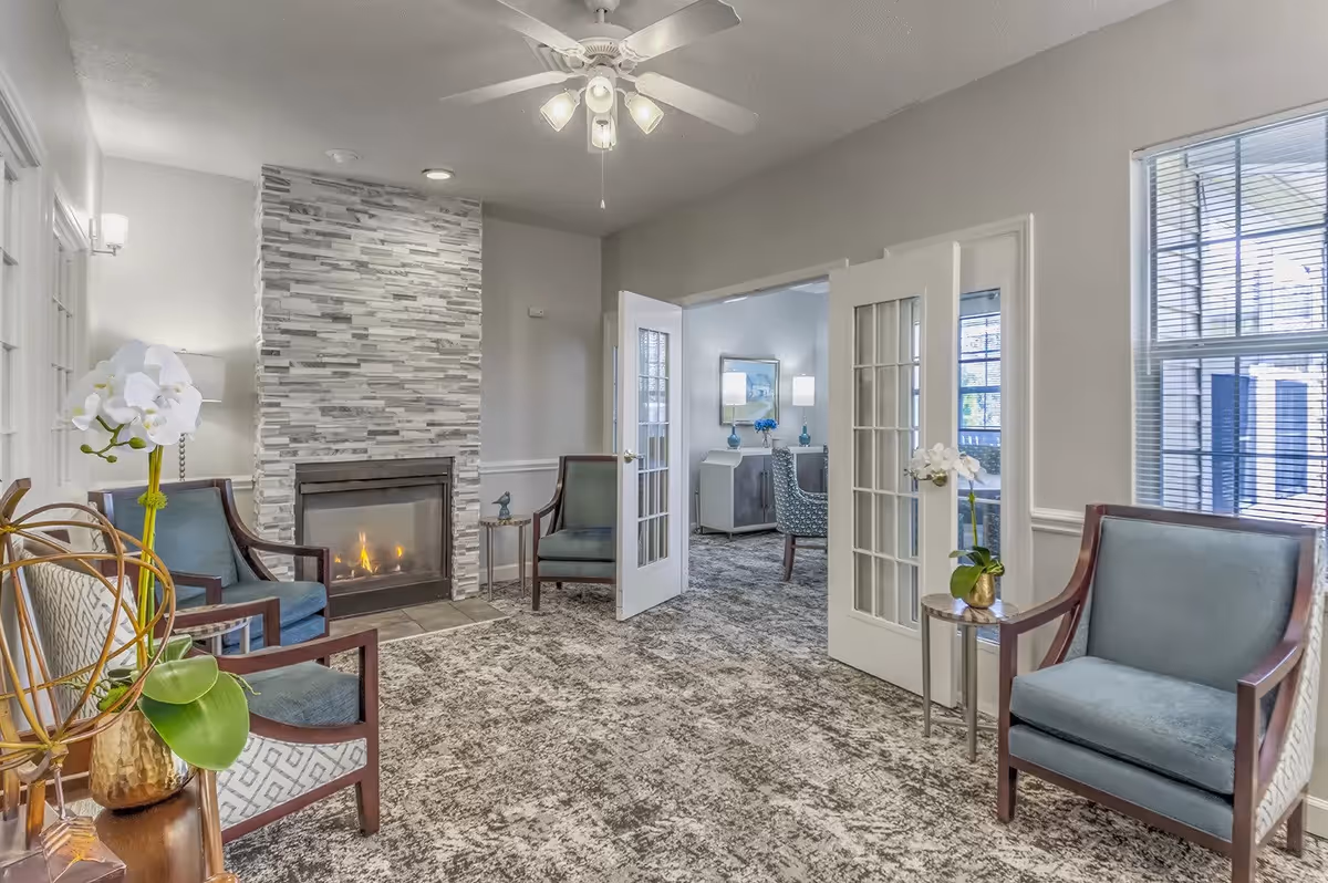 A cozy living room area with a modern stone fireplace, four blue upholstered armchairs, a ceiling fan with lights, and a carpeted floor. The room has white walls and large windows with blinds. French doors open to another room with additional seating and decor.