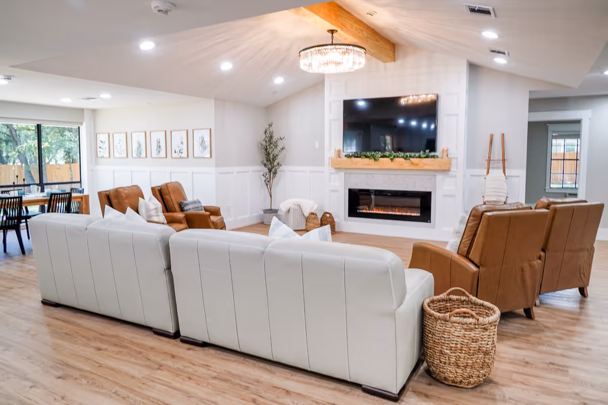 A spacious and bright living room area with white leather sofas and brown leather armchairs arranged around a modern electric fireplace with a mounted flat-screen TV above it. The room features light wood flooring, a large window with a view of greenery outside, framed botanical artwork on the wall, a woven basket, and a ceiling light fixture with a wooden beam accent.