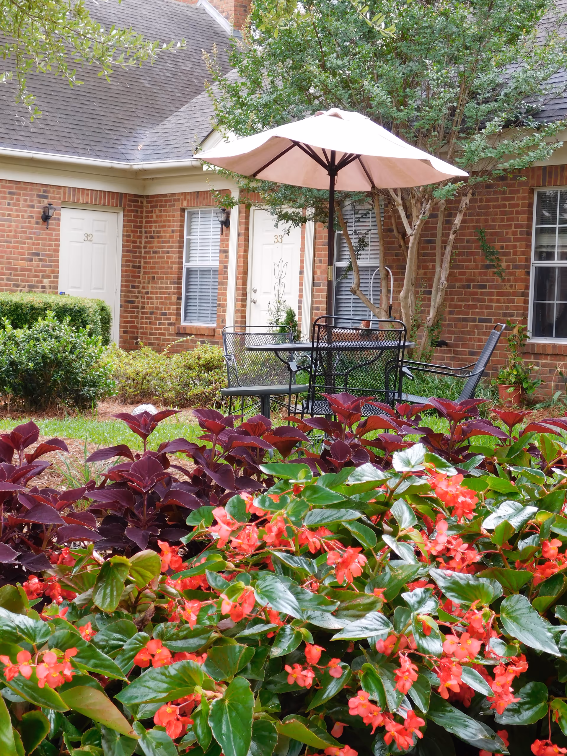 Outdoor patio area with a black metal table and chairs under a large beige umbrella, surrounded by vibrant red and purple flowering plants and greenery. In the background, there is a brick building with white doors numbered 32 and 33 and windows with white blinds.