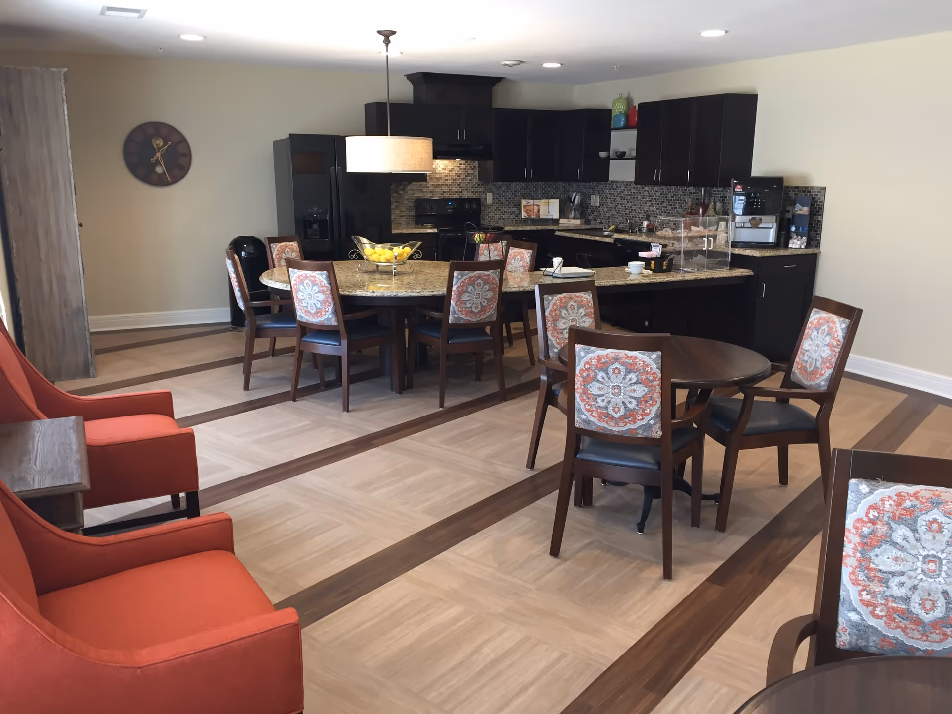 A spacious dining area with a kitchen in the background. The room features a large round dining table with eight chairs that have patterned cushions, a smaller round table with four matching chairs, and two orange armchairs. The kitchen has dark cabinets, a tiled backsplash, and various appliances including a refrigerator, stove, and coffee machine. The floor has a light wood pattern with darker wood inlays.