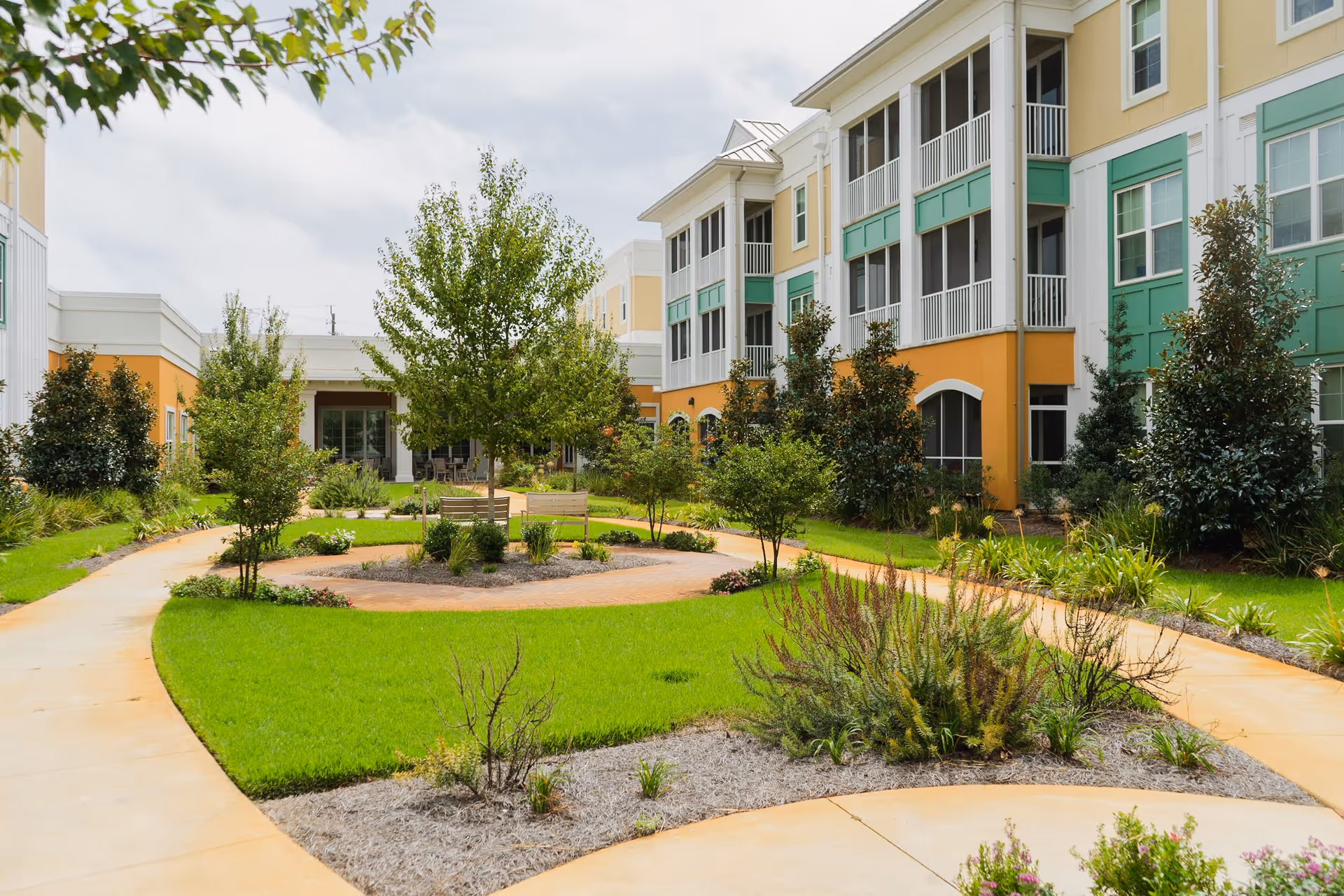 Outdoor courtyard area at The Arbors of Gulf Breeze featuring a circular walking path, green grass, various shrubs and trees, benches, and a multi-story building with balconies in the background under a cloudy sky.