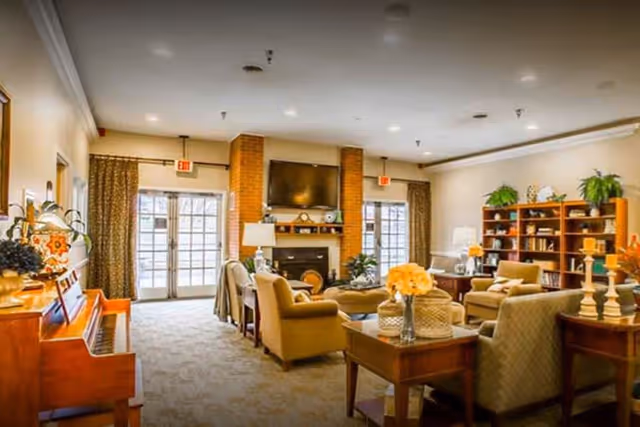 A cozy and well-lit living room area with comfortable armchairs and sofas arranged around a central coffee table. There is a fireplace with a mounted flat-screen TV above it, flanked by two large windows with curtains. A wooden bookshelf filled with books and decorative plants is visible on the right side. A piano is positioned on the left side of the room. The room has a warm and inviting atmosphere with soft lighting and neutral tones.