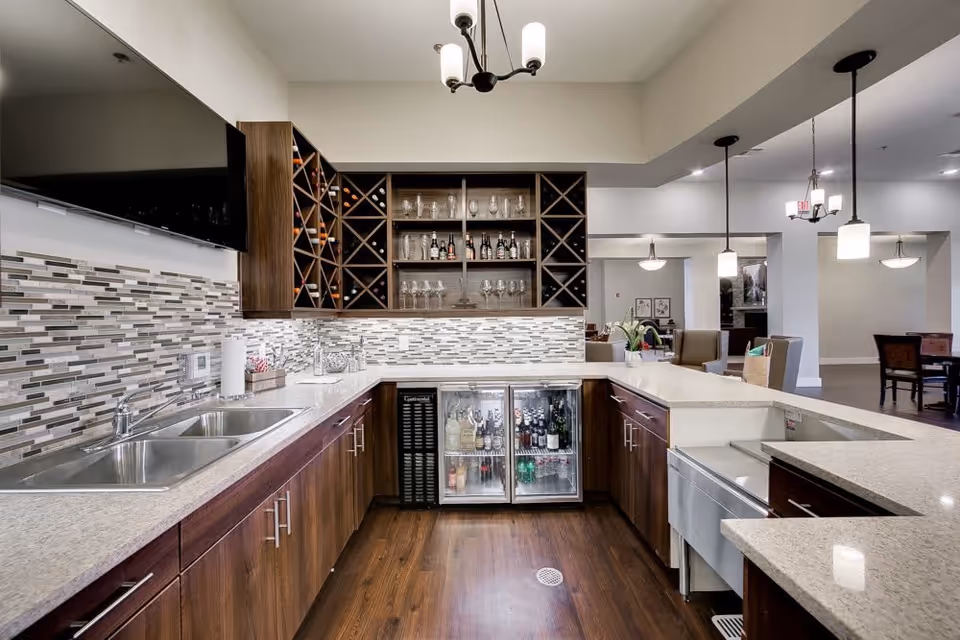 Modern kitchen area with wooden cabinets, a double sink, a tiled backsplash, a wine rack with bottles and glasses, two glass-door refrigerators stocked with beverages, and pendant lighting. The kitchen opens into a dining and living area with tables, chairs, and soft lighting fixtures.