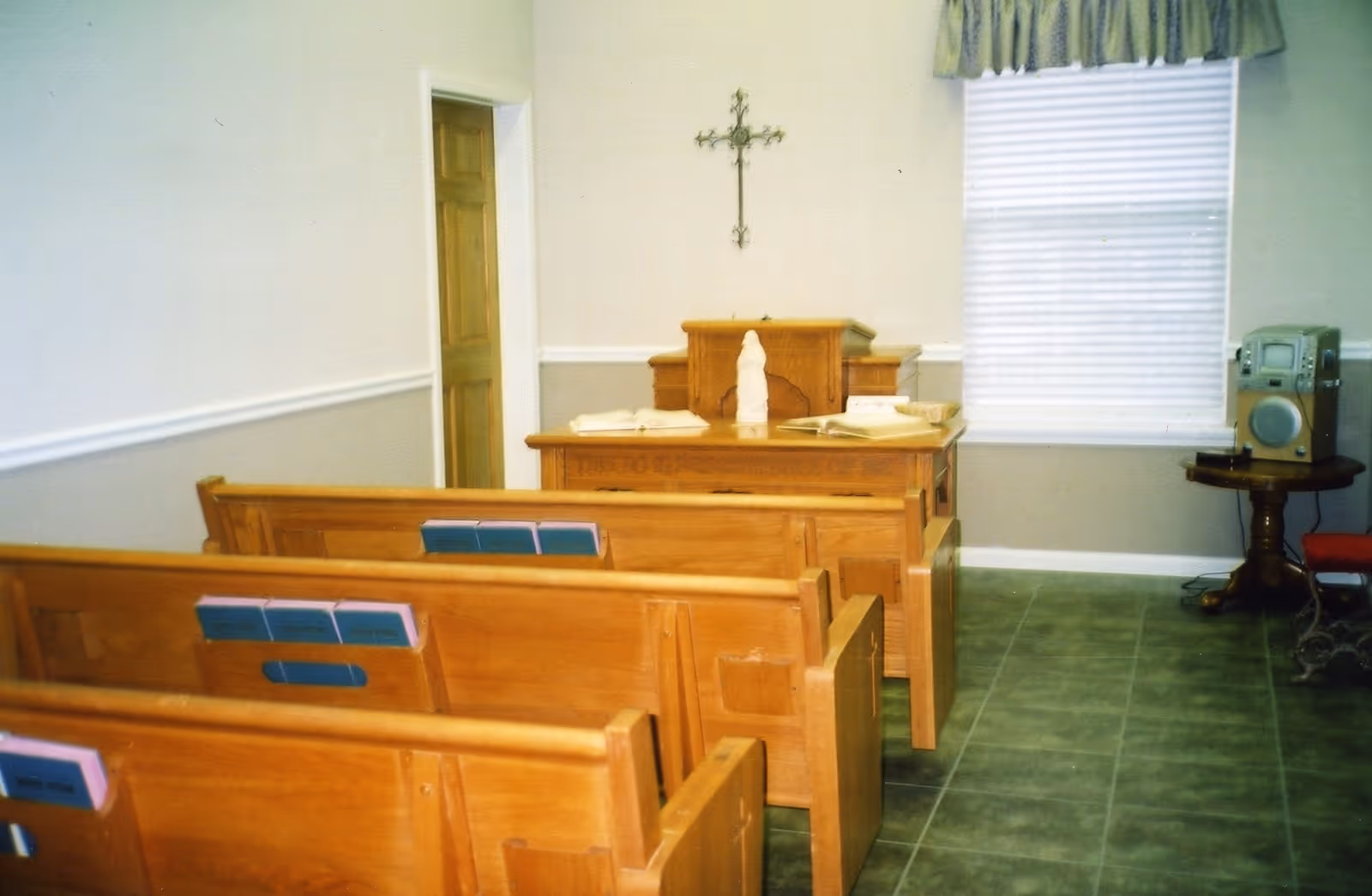 Small chapel room with wooden pews facing a wooden altar. The altar has religious books and a statue on it, with a cross mounted on the wall behind. There is a window with blinds and a valance on the right side, and a small table holding a stereo system.