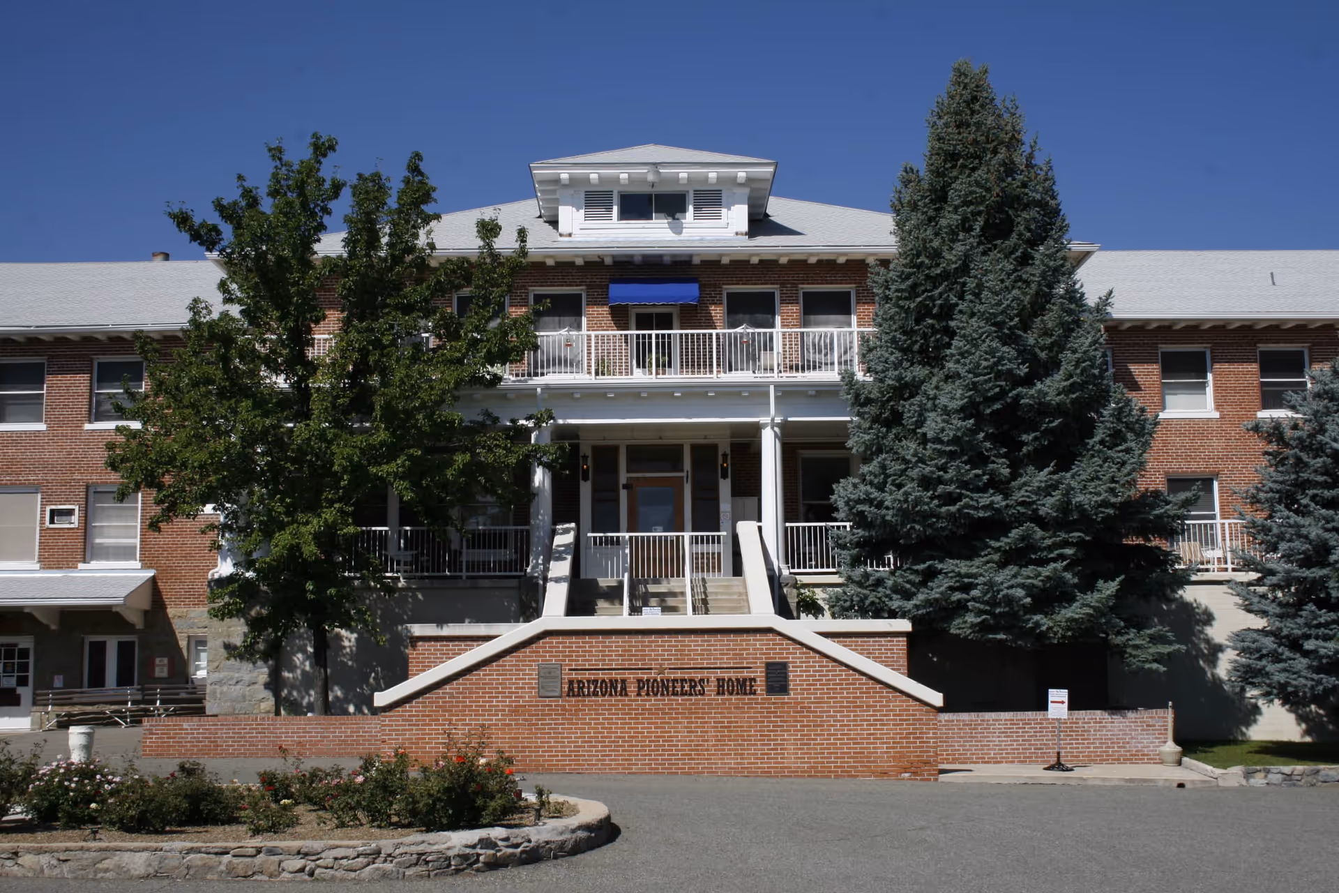 Front exterior view of the Arizona Pioneers' Home, a large brick building with white trim, a central staircase leading to the entrance, and surrounded by trees and landscaping under a clear blue sky.