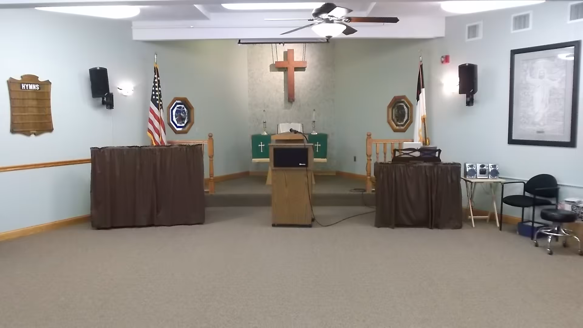 Interior view of a small chapel or worship room with a wooden cross mounted on the back wall above a podium. There are two flags on either side of the podium, an American flag on the left and a Christian flag on the right. The room has light-colored walls, a ceiling fan with lights, and carpeted floor. On the left wall, there is a hymns board, and on the right wall, there is a framed religious picture. Two tables covered with brown cloth are positioned on either side of the podium, with audio equipment on the right table.