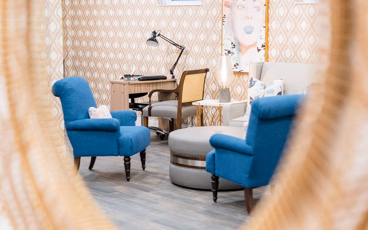 Cozy seating area with two blue armchairs, a round ottoman, a desk and patterned wallpaper.