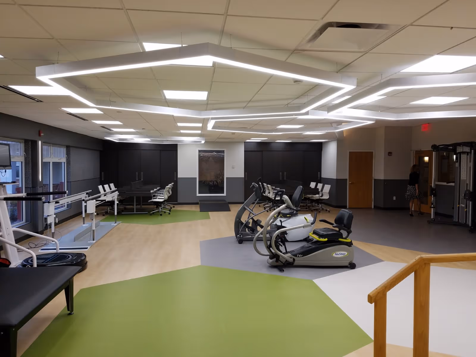A spacious fitness and rehabilitation room with exercise equipment including a NuStep recumbent cross trainer, parallel bars for walking support, and various chairs and tables along the walls. The room has modern hexagonal ceiling lights and a mix of green, gray, and beige flooring. A woman is seen walking near the back door.