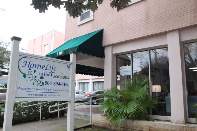 Exterior view of HomeLife in the Gardens, LLC facility showing a building entrance with a green awning displaying the number 1101. A white sign in front of the building displays the facility name, phone number, services offered, and website. There are some plants and parked cars visible in the background.