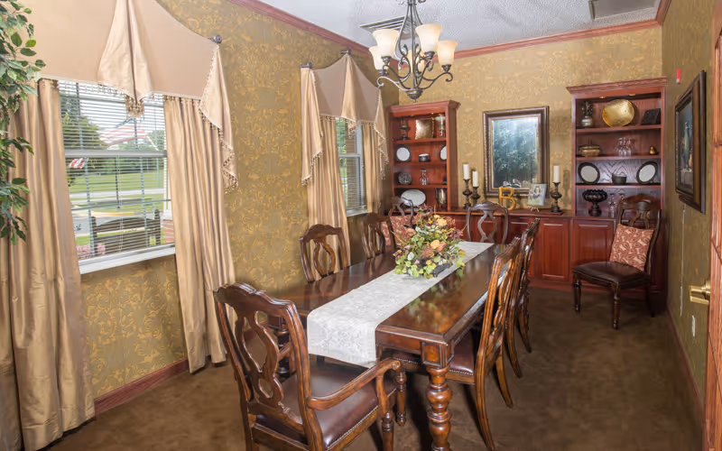A formal dining room with a long wooden table and eight matching chairs. The table is decorated with a white table runner and a floral centerpiece. The room has gold patterned wallpaper, two windows with beige curtains, a chandelier hanging from the ceiling, and built-in wooden shelves and cabinets along the far wall displaying decorative items.
