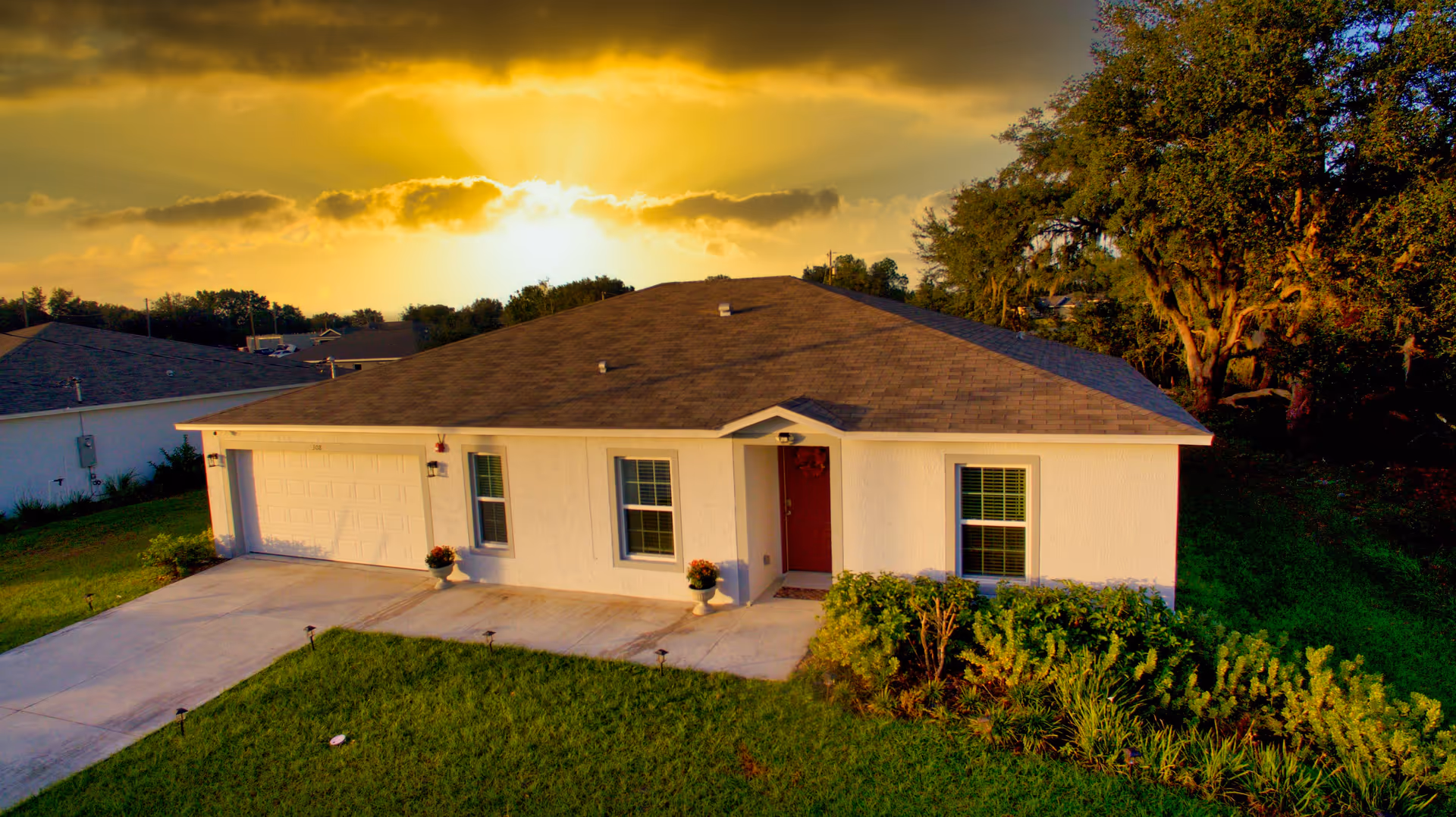 Single-story house with a brown shingled roof, white exterior walls, a red front door, and two windows with blinds. The house has a two-car garage and a concrete driveway. There are green bushes and a well-maintained lawn in front of the house. The sun is setting in the background, casting a warm golden light over the scene.