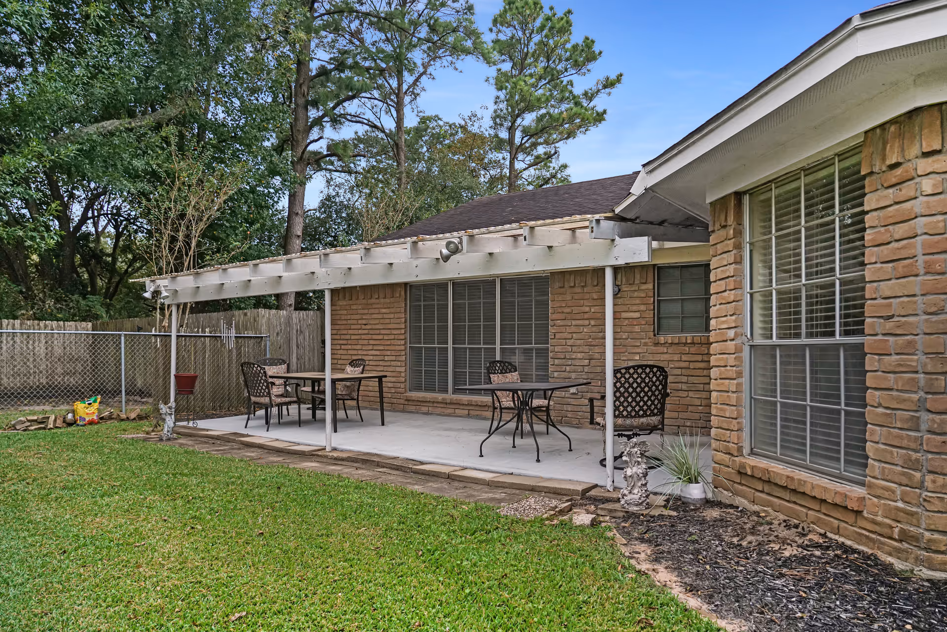 Outdoor patio area with a white pergola attached to a brick building. The patio has two tables and several chairs on a concrete floor. There is a grassy lawn in the foreground and trees and a wooden fence in the background.