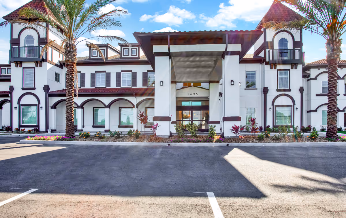 Front exterior view of The Blake at LPGA facility with a white building featuring dark brown trim, large entrance canopy, palm trees, and a parking area in front under a partly cloudy sky.